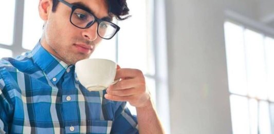 Hombre joven con lentes mira su móvil mientras toma café