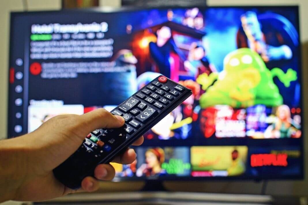 Fotografía de un joven que tiene un control remoto en la mano y en el fondo un televisor con imágenes